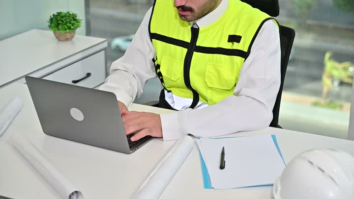 العمل على الحاسوب المحمول، الاهتمام بالتطور العمراني بسواعد المهندسين السعوديين, مهندس عربي خليجي سعودي يرتدي سترة العمل وخوذة الحماية البيضاء ينجز المهام اليومية في المكتب ، مفهوم الهندسة والإنشاءات المعمارية، مشاريع إعمارية سعودية وطنية