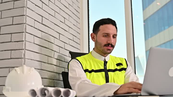 Working in the engineering sector, Saudi national construction projects, working on a laptop, taking care of urban development with the help of Saudi engineers, a Saudi Arabian Gulf engineer wearing a work jacket and a white protective helmet performing daily tasks in the office, the concept of engineering and architectural construction.