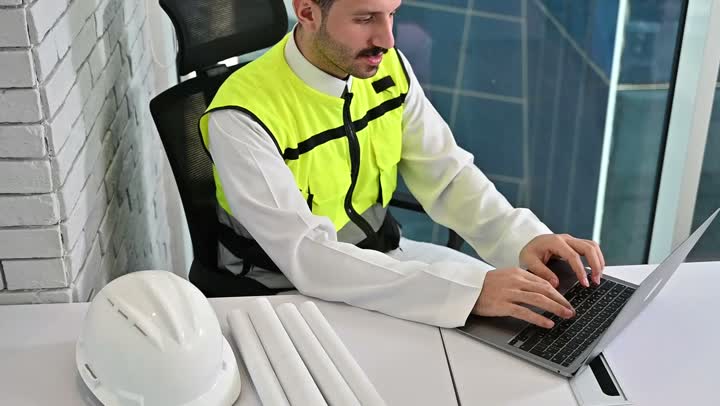 العمل في القطاع الهندسي، مشاريع إعمارية سعودية وطنية، العمل على الحاسوب المحمول، الاهتمام بالتطور العمراني بسواعد المهندسين السعوديين, مهندس عربي خليجي سعودي يرتدي سترة العمل وخوذة الحماية البيضاء ينجز المهام اليومية في المكتب، مفهوم الهندسة والإنشاءات المعمارية