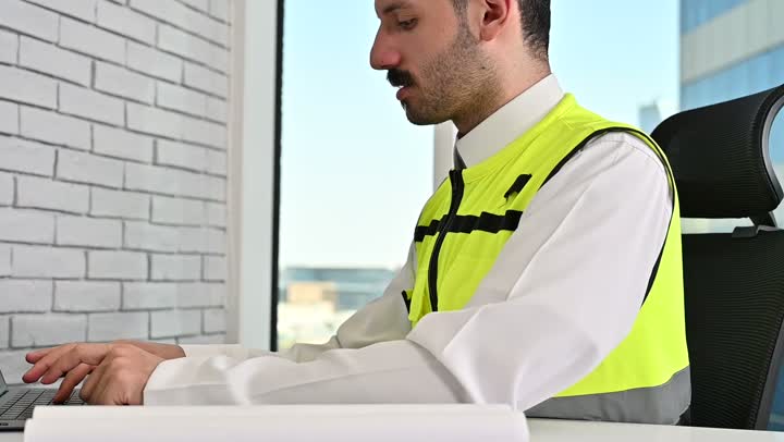العمل في القطاع الهندسي، مشاريع إعمارية سعودية وطنية، العمل على الحاسوب المحمول، الاهتمام بالتطور العمراني بسواعد المهندسين السعوديين, مهندس عربي خليجي سعودي يرتدي سترة العمل وخوذة الحماية البيضاء ينجز المهام اليومية في المكتب، مفهوم الهندسة والإنشاءات المعمارية