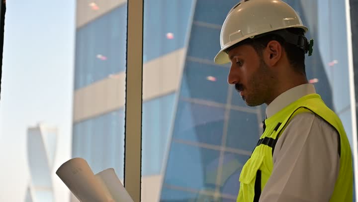 متابعة مخطط المشروع الهندسي وتدقيقه, مفهوم الهندسة والإنشاءات المعمارية، مشاريع إعمارية سعودية وطنية، الاهتمام بالتطور العمراني بسواعد المهندسين السعوديين, مهندس عربي خليجي سعودي يرتدي سترة العمل وخوذة الحماية البيضاء ينجز المهام اليومية في المكتب