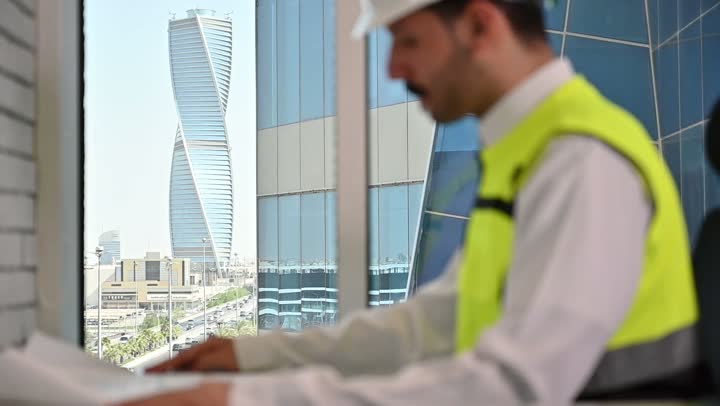 متابعة مخطط المشروع الهندسي وتدقيقه, مفهوم الهندسة والإنشاءات المعمارية، مشاريع إعمارية سعودية وطنية، الاهتمام بالتطور العمراني بسواعد المهندسين السعوديين, مهندس عربي خليجي سعودي يرتدي سترة العمل وخوذة الحماية البيضاء ينجز المهام اليومية في المكتب