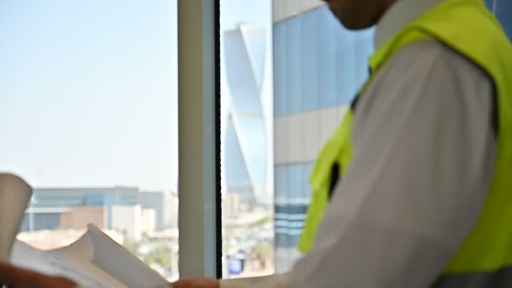 الاهتمام بالتطور العمراني بسواعد المهندسين السعوديين, مهندس عربي خليجي سعودي يرتدي سترة العمل وخوذة الحماية البيضاء ينجز المهام اليومية في المكتب، متابعة مخطط المشروع الهندسي وتدقيقه, مفهوم الهندسة والإنشاءات المعمارية، مشاريع إعمارية سعودية وطنية