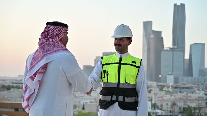 التفاهم والتعاون بين الزملاء، تبادل الخبرات والمعلومات لتحقيق الاهداف، مهندس عربي خليجي سعودي يرتدي سترة العمل وخوذة الحماية البيضاء يقف مع زميله، متابعة مخطط المشروع الهندسي وتدقيقه, مفهوم الهندسة والإنشاءات المعمارية، مشاريع إعمارية سعودية وطنية