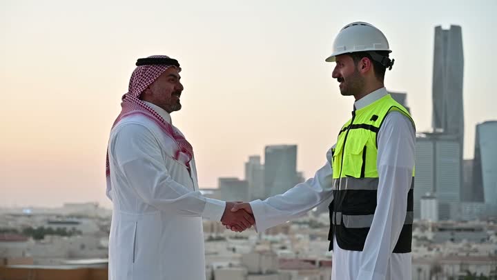 التفاهم والتعاون بين الزملاء، تبادل الخبرات والمعلومات لتحقيق الاهداف، مهندس عربي خليجي سعودي يرتدي سترة العمل وخوذة الحماية البيضاء يقف مع زميله، متابعة مخطط المشروع الهندسي وتدقيقه, مفهوم الهندسة والإنشاءات المعمارية، مشاريع إعمارية سعودية وطنية