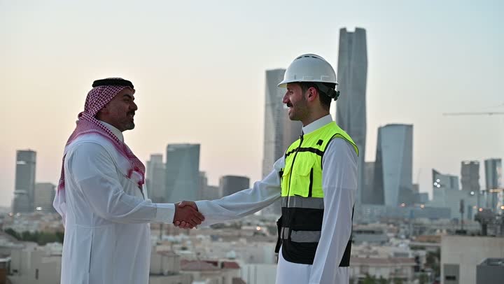 التفاهم والتعاون بين الزملاء، تبادل الخبرات والمعلومات لتحقيق الاهداف، مهندس عربي خليجي سعودي يرتدي سترة العمل وخوذة الحماية البيضاء يقف مع زميله، متابعة مخطط المشروع الهندسي وتدقيقه, مفهوم الهندسة والإنشاءات المعمارية، مشاريع إعمارية سعودية وطنية