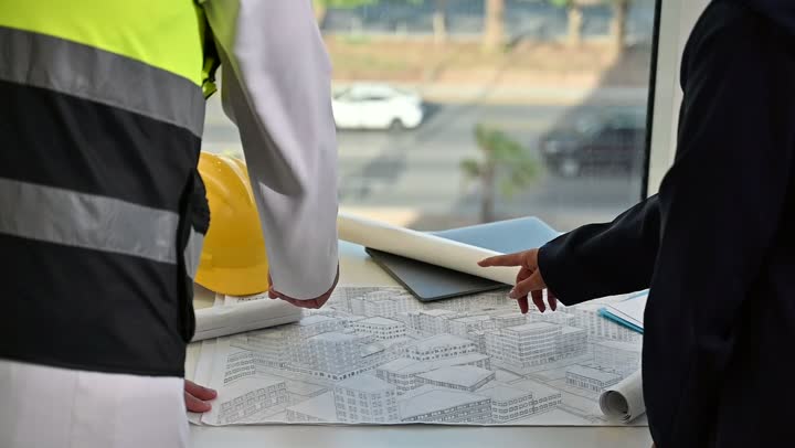 Exchanging experiences and information to achieve goals. A Saudi Arabian Gulf engineer wearing a work jacket and a white protective helmet stands with his colleague, following up and reviewing the engineering project plan. The concept of engineering and architectural construction, Saudi national construction projects, understanding and cooperation between colleagues.