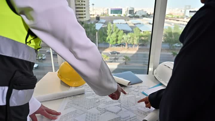 تبادل الخبرات والمعلومات لتحقيق الاهداف، مهندس عربي خليجي سعودي يرتدي سترة العمل وخوذة الحماية البيضاء يقف مع زميلته، متابعة مخطط المشروع الهندسي وتدقيقه, مفهوم الهندسة والإنشاءات المعمارية، مشاريع إعمارية سعودية وطنية، التفاهم والتعاون بين الزملاء