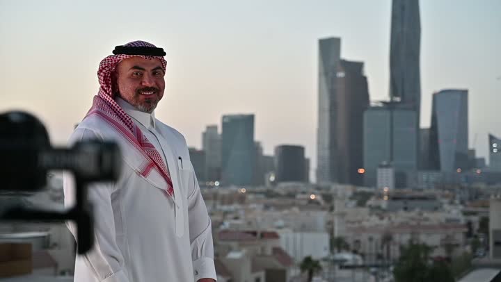 استخدام كاميرا رقمية عالية الدقة، إتقان واحتراف مهنة التصوير، الاهتمام بالمظهر الخارجي، مفهوم الرجولة والأناقة، مصور فوتوغرافي يقوم بتصوير رجل عربي خليجي سعودي يرتدي الثوب التقليدي والشماغ يقف بإيماءات الفرح والسرور، خلفية الابراج وناطحات السحاب في الرياض