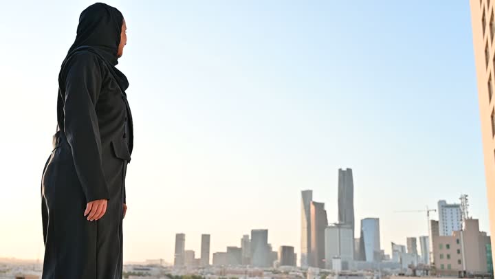 الثقة والاعتزاز بالنفس، الاهتمام بالأناقة والمظهر الخارجي، مفهوم الأنوثة والنعومة، امرأة عربية خليجية سعودية مبتسمة محجبة ترتدي عباءة تقف بإيماءات السرور والسعادة، خلفية الابراج وناطحات السحاب في الرياض
