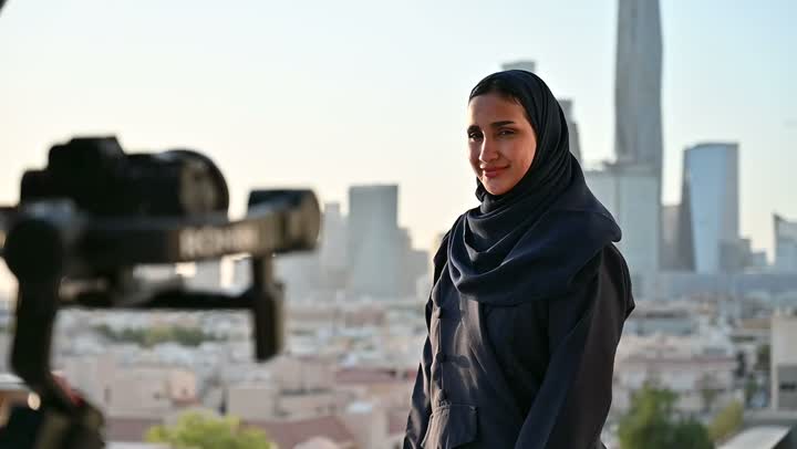 إتقان واحتراف مهنة التصوير، استخدام كاميرا رقمية عالية الدقة، الاهتمام بالأناقة والمظهر الخارجي، مفهوم الأنوثة والنعومة، مصور يقوم بتصوير امرأة عربية خليجية سعودية مبتسمة محجبة ترتدي عباءة تقف بإيماءات السرور والسعادة، خلفية الابراج وناطحات السحاب في الرياض
