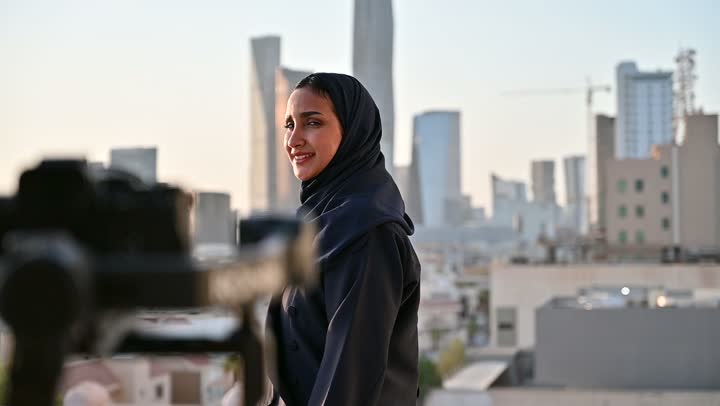 إتقان واحتراف مهنة التصوير، استخدام كاميرا رقمية عالية الدقة، الاهتمام بالأناقة والمظهر الخارجي، مفهوم الأنوثة والنعومة، مصور يقوم بتصوير امرأة عربية خليجية سعودية مبتسمة محجبة ترتدي عباءة تقف بإيماءات السرور والسعادة، خلفية الابراج وناطحات السحاب في الرياض