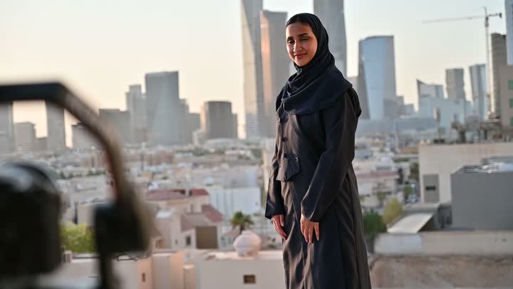 إتقان واحتراف مهنة التصوير، استخدام كاميرا رقمية عالية الدقة، الاهتمام بالأناقة والمظهر الخارجي، مفهوم الأنوثة والنعومة، مصور يقوم بتصوير امرأة عربية خليجية سعودية مبتسمة محجبة ترتدي عباءة تقف بإيماءات السرور والسعادة، خلفية الابراج وناطحات السحاب في الرياض