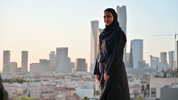 إتقان واحتراف مهنة التصوير، استخدام كاميرا رقمية عالية الدقة، الاهتمام بالأناقة والمظهر الخارجي، مفهوم الأنوثة والنعومة، مصور يقوم بتصوير امرأة عربية خليجية سعودية مبتسمة محجبة ترتدي عباءة تقف بإيماءات السرور والسعادة، خلفية الابراج وناطحات السحاب في الرياض