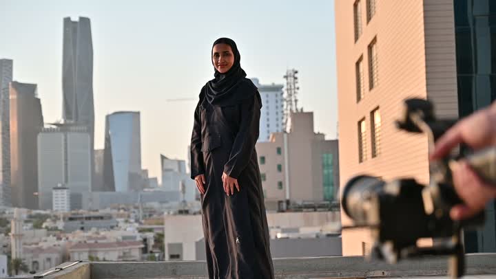 إتقان واحتراف مهنة التصوير، استخدام كاميرا رقمية عالية الدقة، الاهتمام بالأناقة والمظهر الخارجي، مفهوم الأنوثة والنعومة، مصور يقوم بتصوير امرأة عربية خليجية سعودية مبتسمة محجبة ترتدي عباءة تقف بإيماءات السرور والسعادة، خلفية الابراج وناطحات السحاب في الرياض