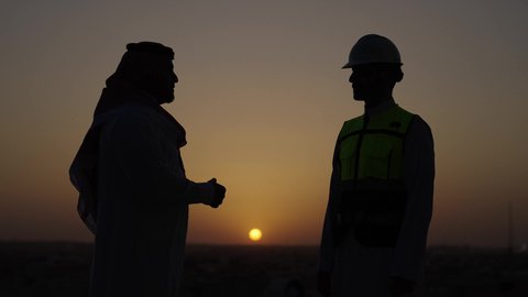 Standing outdoors, agreement and partnership on business and projects, new successful business deals between businessmen, side view of a Saudi Arabian Gulf man wearing a thobe and shemagh shaking hands with an engineer wearing a helmet and a safety vest, sunset background