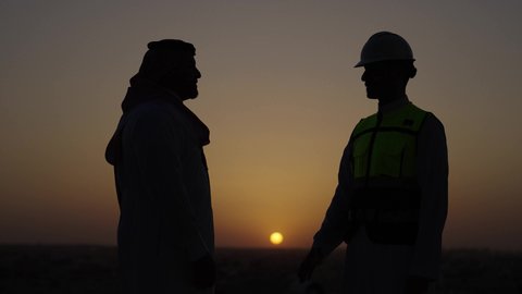 الوقوف في الهواء الطلق، الاتفاق والشراكة على الأعمال والمشاريع، صفقات عمل جديدة ناجحة بين رجال الأعمال، تصوير من الجانب لرجل عربي خليجي سعودي يرتدي الثوب والشماغ يقوم بمصافحة مهندس يرتدي الخوذة وسترة الحماية، خلفية غروب الشمس