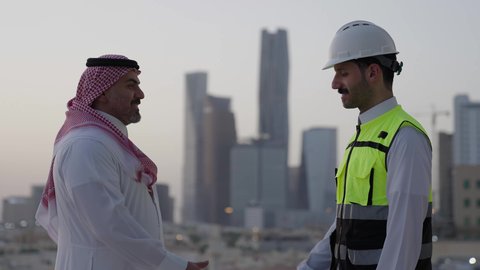ايماءات السعادة والسرور، الوقوف في الهواء الطلق، الاتفاق والشراكة على الأعمال والمشاريع، عقد صفقات عمل جديدة، تصوير من الجانب لرجل عربي خليجي سعودي يرتدي الثوب والشماغ يقوم بمصافحة مهندس يرتدي الخوذة وسترة الحماية، خلفية الابراج وناطحات السحاب
