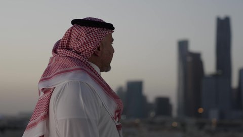 التقدم والتطور المعماري في العاصمة، الوقوف في الهواء الطلق، تصوير مقرب من الجانب لرجل عربي خليجي سعودي يرتدي الثوب والشماغ التقليدي، خلفية الابراج وناطحات السحاب في الرياض