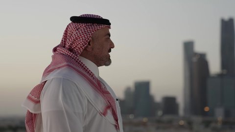 التقدم والتطور المعماري في العاصمة، الوقوف في الهواء الطلق، تصوير مقرب من الجانب لرجل عربي خليجي سعودي يرتدي الثوب والشماغ التقليدي، خلفية الابراج وناطحات السحاب في الرياض