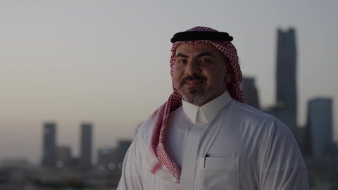 الوقوف بثقة والنظر إلى الكاميرا بإيماءات السرور، التقدم والتطور المعماري في العاصمة، الوقوف في الهواء الطلق، تصوير مقرب لرجل عربي خليجي سعودي يرتدي الثوب والشماغ التقليدي، خلفية الابراج وناطحات السحاب في الرياض