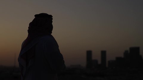 الوقوف بثقة وتأمل المدينة، الوقوف في الهواء الطلق، تصوير مقرب من الجانب لرجل عربي خليجي سعودي يرتدي الثوب والشماغ التقليدي، خلفية غروب الشمس