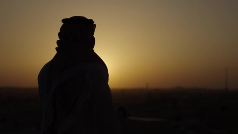 الوقوف بثقة وتأمل معالم المدينة، الخروج في الهواء الطلق، تصوير من الجانب لرجل عربي خليجي سعودي يرتدي الثوب والشماغ التقليدي، خلفية غروب الشمس