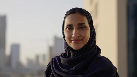 Architectural progress and development in the Gulf States, a background of towers and skyscrapers in Riyadh, a close-up of a smiling veiled Saudi Arabian Gulf woman looking at the camera with gestures of pleasure and happiness, standing proudly in honor of the Saudi state.