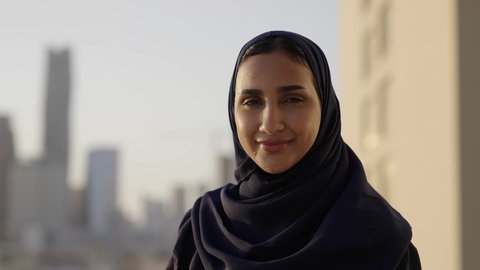 التقدم والتطور المعماري في دول الخليج، خلفية أبراج وناطحات سحاب مدينة الرياض، تصوير مقرب لامرأة عربية خليجية سعودية محجبة مبتسمة تنظر إلى الكاميرا بإيماءات السرور والسعادة، الوقوف بفخر واعتزاز بالدولة السعودية.