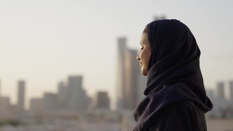 الوقوف بفخر واعتزاز بالدولة السعودية، التقدم والتطور المعماري في دول الخليج، خلفية أبراج وناطحات سحاب مدينة الرياض، تصوير مقرب من الجانب لامرأة عربية خليجية سعودية محجبة تقوم بتأمل معالم المدينة