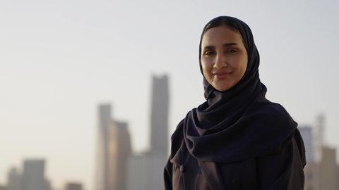 التقدم والتطور المعماري في دول الخليج، خلفية أبراج وناطحات سحاب مدينة الرياض، تصوير مقرب لامرأة عربية خليجية سعودية محجبة مبتسمة تنظر إلى الكاميرا بإيماءات السرور والسعادة، الوقوف بفخر واعتزاز بالدولة السعودية.
