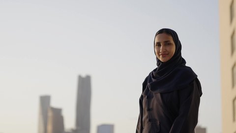 خلفية أبراج وناطحات سحاب مدينة الرياض، امرأة عربية خليجية سعودية محجبة مبتسمة تنظر إلى الكاميرا بإيماءات السرور والسعادة، التقدم والتطور المعماري في دول الخليج، الوقوف بفخر واعتزاز بالدولة السعودية.