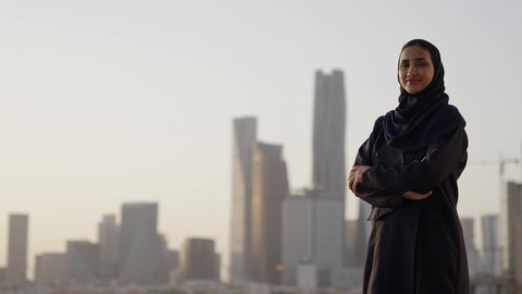 Standing proudly in honor of the Saudi state, the progress and architectural development in the Gulf states, a background of towers and skyscrapers in Riyadh, a veiled Saudi Arabian Gulf woman standing with crossed hands and looking at the camera with gestures of self-confidence
