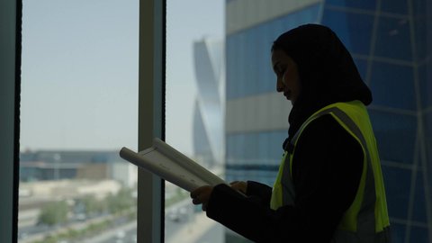 العمل في المجال الهندسي، مهن ووظائف مكتبية نسائية، مفهوم الهندسة والإنشاءات المعمارية، مهندسة عربية خليجية سعودية محجبة ترتدي سترة الحماية تقراء المخطط الهندسي وتنظر من خلال النافذة، خلفية أبراج وناطحات السحاب