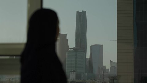 تأمل أبراج وناطحات سحاب مدينة الرياض، تصوير مقرب من الجانب لامرأة عربية خليجية سعودية محجبة تقف داخل المبنى، التقدم والتطور المعماري في دول الخليج، الوقوف بفخر واعتزاز بالدولة السعودية.