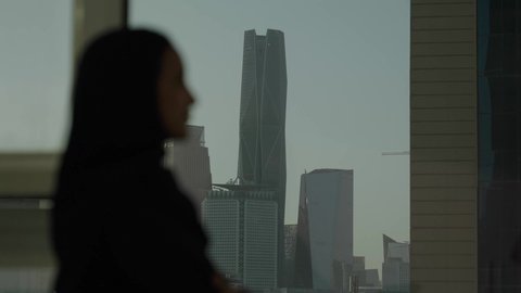 تأمل أبراج وناطحات سحاب مدينة الرياض، تصوير مقرب من الجانب لامرأة عربية خليجية سعودية محجبة تقف داخل المبنى، التقدم والتطور المعماري في دول الخليج، الوقوف بفخر واعتزاز بالدولة السعودية.