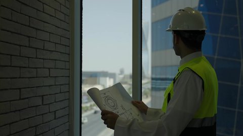 متابعة استراتيجيات المشروع، العمل في المجال الهندسي، مفهوم الهندسة والإنشاءات المعمارية، مهندس عربي خليجي سعودي يرتدي سترة و خوذة الحماية يقراء المخطط الهندسي وينظر من خلال النافذة، خلفية أبراج وناطحات السحاب، استخدام معدات العمل