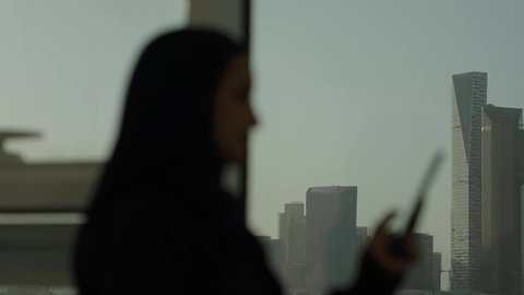 جهاز تقني حديث ومتطور، أبراج وناطحات سحاب مدينة الرياض، تصوير من الجانب لامرأة عربية خليجية سعودية محجبة تقف داخل المبنى تقوم باستخدام الهاتف المحمول، التقدم والتطور المعماري في دول الخليج