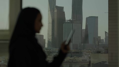 جهاز تقني حديث ومتطور، أبراج وناطحات سحاب مدينة الرياض، تصوير من الجانب لامرأة عربية خليجية سعودية محجبة تقف داخل المبنى تقوم باستخدام الهاتف المحمول، التقدم والتطور المعماري في دول الخليج
