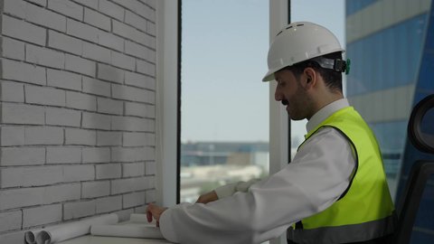 تدقيق ومتابعة استراتيجيات المشروع، العمل في المجال الهندسي، مفهوم الهندسة والإنشاءات المعمارية، مهندس عربي خليجي سعودي يرتدي  الخوذة وسترة العمل يقراء المخطط الهندسي، خلفية أبراج وناطحات السحاب، استخدام معدات العمل، مهن ووظائف سعودية