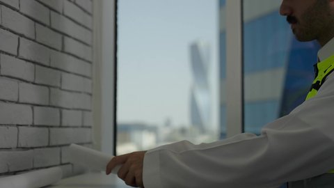 الأشراف على العمل، العمل في المجال الهندسي، مفهوم الهندسة والإنشاءات المعمارية، تصوير مقرب لمهندس عربي خليجي سعودي يرتدي  الخوذة وسترة العمل يقراء المخطط الهندسي، خلفية أبراج وناطحات السحاب، استخدام معدات العمل، مهن ووظائف سعودية