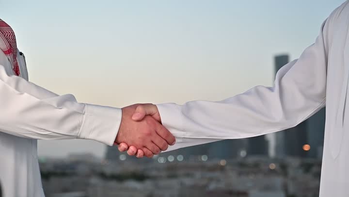الاتفاق والشراكة على الأعمال والمشاريع التجارية، تعارف وصداقات وبناء علاقات اجتماعية، صفقات عمل جديدة ناجحة بين رجال الأعمال، الخروج في الهواء الطلق، رجلان عربيان خليجيان سعوديان يقومان بمصافحة بعضهما، خلفية الابراج وناطحات السحاب في الرياض
