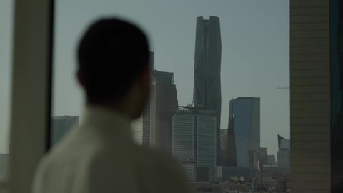 أبراج وناطحات سحاب مدينة الرياض، تصوير مقرب من الجانب لرجل عربي خليجي سعودي يرتدي الثوب التقليدي يقف داخل المبنى يقوم بتأمل معالم المدينة، التقدم والتطور المعماري في دول الخليج