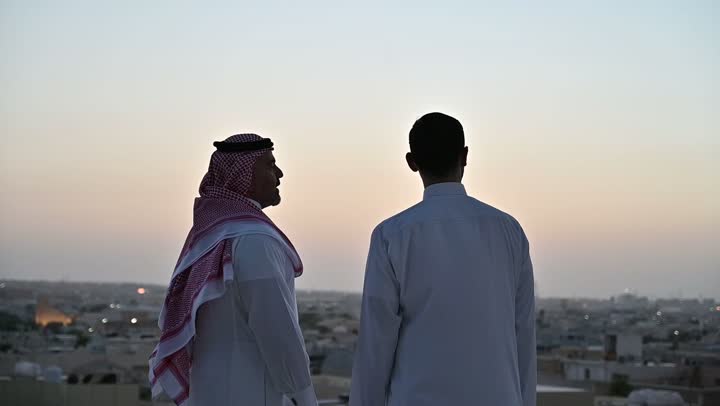 تبادل الأحاديث والاخبار بين الاصدقاء، الاهتمام بالمظهر الخارجي، مفهوم الرجولة والأناقة، رجل عربي خليجي سعودي يرتدي الثوب التقليدي والشماغ يقف مع صديقه بإيماءات الفرح والسرور، خلفية الابراج وناطحات السحاب في الرياض