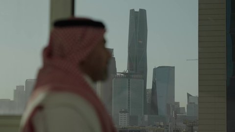 الفخر والاعتزاز بالدولة السعودية، أبراج وناطحات سحاب مدينة الرياض، تصوير مقرب من الجانب لرجل عربي خليجي سعودي يرتدي الثوب والشماغ التقليدي يقف داخل المبنى يقوم بتأمل معالم المدينة، التقدم والتطور المعماري في دول الخليج