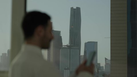  جهاز تقني حديث ومتطور،  تصوير مقرب من الجانب لرجل عربي خليجي سعودي يرتدي الثوب التقليدي يقف داخل المبنى يقوم باستخدام الهاتف المحمول، التقدم والتطور المعماري في دول الخليج، خلفية الأبراج وناطحات السحاب المالية في مدينة الرياض