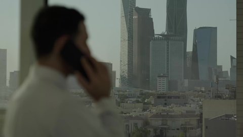  استخدام جهاز تقني حديث ومتطور،  تصوير مقرب من الجانب لرجل عربي خليجي سعودي يرتدي الثوب التقليدي يقف داخل المبنى يقوم بإجراء مكالمة هاتفية عبر الهاتف المحمول، التقدم والتطور المعماري في دول الخليج، خلفية الأبراج وناطحات السحاب المالية في مدينة الرياض