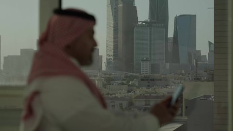  جهاز تقني حديث ومتطور،  تصوير مقرب من الجانب لرجل عربي خليجي سعودي يرتدي الثوب والشماغ التقليدي يقف داخل المبنى يقوم باستخدام الهاتف المحمول، التقدم والتطور المعماري في دول الخليج، خلفية الأبراج وناطحات السحاب المالية في مدينة الرياض