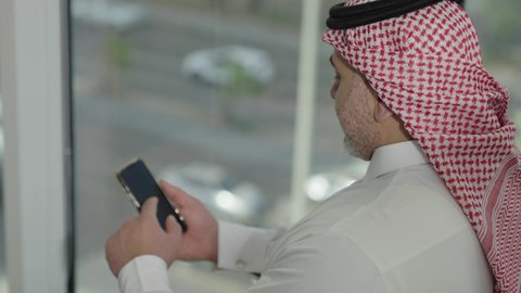  تصفح مواقع التواصل الاجتماعي، جهاز تقني حديث ومتطور،  تصوير مقرب من الجانب لرجل عربي خليجي سعودي يرتدي الثوب والشماغ التقليدي يقف داخل المبنى يقوم باستخدام الهاتف المحمول، إنجاز العمل عن بعد