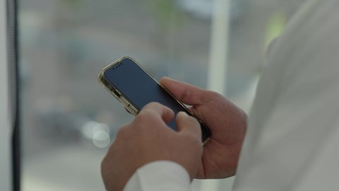 إنجاز العمل عن بعد، تصفح مواقع التواصل الاجتماعي، جهاز تقني حديث ومتطور،  تصوير مقرب من الجانب لرجل عربي خليجي سعودي يرتدي الثوب التقليدي يقف داخل المبنى يقوم باستخدام الهاتف المحمول